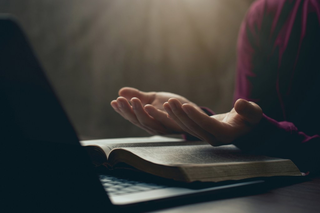 Prayerful open hands over a sacred text in front of a laptop.